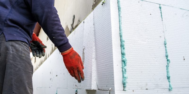 Construction worker installing styrofoam insulation sheets on house facade wall for thermal protection.